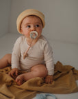 A baby wearing a beige knit hat and cream long-sleeve onesie sits on a brown blanket, looking to the side while using the FRIGG Daisy Natural Rubber Pacifier from FRIGG.