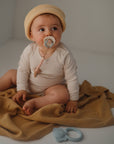 A baby in a cream ribbed onesie and beige knit hat sits on a mustard blanket, holding a pacifier, with the mushie Silicone Pacifier Clip | Eva among other pacifier accessories nearby.