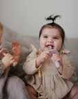 Two smiling babies with pigtails sit on a couch, each happily chewing on a mushie Flower Training Toothbrush—one in a beige dress, the other in grey.