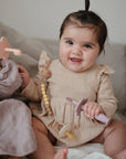 A smiling baby with dark hair in a topknot sits on a couch, wearing a beige dress and holding the mushie Silicone Pacifier Clip | Eva. Another baby in a gray outfit is partially visible beside her.