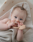 A light-haired baby wrapped in a beige towel lies on a blanket, gazing up while holding the mushie Dog Teether, with some cream visible on their cheek.