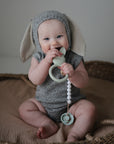A baby in a gray knit bunny outfit sits on a brown blanket in a woven basket, smiling and holding a green pacifier with the mushie Silicone Pacifier Clip | Hera.