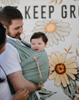 A smiling baby is carried in a green mushie Baby Wrap made from organic cotton by an adult with brown hair and beard, while another person stands nearby. They are in front of a large floral mural with partially visible text.