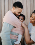 A man carries a baby in a pink wrap carrier with the mushie Silicone Pacifier Clip | Eva, made from food-grade silicone, hanging from the wrap. A smiling woman stands beside them adjusting the carrier; vertical wooden panels are in the background.