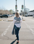 A woman in jeans and a white shirt crosses a street on a sunny day, carrying her baby in the mushie Baby Wrap, with green traffic lights behind her.
