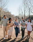 A group of six adults, three carrying babies in breathable mushie Baby Wraps, walk outdoors on a sunny day, smiling and talking amid bare trees and string lights.
