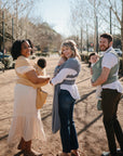 Three adults, each carrying a baby in a mushie Baby Wrap, smile at the camera outdoors on a sunny day. Trees, parked cars, and a dirt path appear in the background.