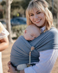 A smiling blonde woman holds a sleeping baby wrapped in a blue carrier outdoors on a sunny day. Nearby, the mushie Silicone Pacifier Clip | Eva, a handy pacifier accessory, is attached for added convenience.