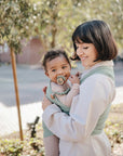 A woman carries a baby in a green wrap outdoors. The baby, wearing the mushie Silicone Pacifier Clip | Hera, looks at the camera as the woman gazes at them. Trees and a paved path form the minimalist backdrop.