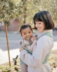 A woman smiles while holding a baby outdoors in a light green wrap. The baby, wearing cozy clothes, uses a FRIGG Daisy Natural Rubber Pacifier from FRIGG as sunlight filters through the trees.