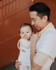 An adult with glasses carries a baby in a white wrap. The baby, dressed in light clothes and holding a FRIGG Moon Natural Rubber Pacifier by FRIGG, looks at the camera. An orange wall with diagonal lines is in the background.