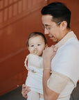 A man wearing glasses holds a baby with a mushie Silicone Pacifier Clip | Luna attached, wrapped in white. The man appears slightly unsure as the baby looks at the camera. A brown staircase can be seen in the background.