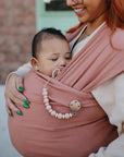 A smiling woman with green nails carries a sleeping baby in a pink wrap. The baby has the mushie Silicone Pacifier Clip | Luna attached—a practical and stylish accessory—set against a softly blurred background.