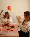 A woman holds a baby with a mushie Silicone Pacifier Clip | Hera at a decorated table with two kids, surrounded by fruits, bowls, plates, and lit candles. Orange paper lanterns above give the setting a warm, cozy ambiance.