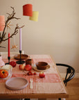 A dining table set with Mushie Classic Silicone Suction Plates, ceramic bowls, apples, and tomatoes on a checkered tablecloth. Red and pink candles, lanterns, and a branch with a gold star hang above as decoration.