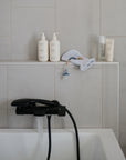 A bathroom scene features a white tub with water running from a black faucet. On the ledge are toiletries, skincare bottles, and the mushie Silicone Pacifier Clip | Eva with a baby pacifier, along with a white washcloth.