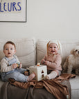 Two babies play with a shape sorter toy on a beige couch, one in overalls and one in a pink outfit with a bonnet. A small white dog sits beside them. The Mushie Ribbed Baby Blanket is draped in the background, along with a framed FLEURS print.