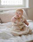 A baby in a beige outfit and knitted bonnet sits on a bed with white bedding, smiling at the camera with one hand in their mouth. The mushie Silicone Pacifier Clip | Hera lies nearby, adding to the minimalist look in soft natural light.