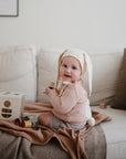 A smiling baby wearing a bunny ear hat and pink sweater sits on a couch next to the Mushie Ribbed Baby Blanket, playing with wooden blocks and looking at the camera.