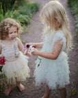 Two young girls in pastel, ruffled dresses stand barefoot on a garden path holding colorful flowers. One, with curly hair, uses the mushie Silicone Pacifier Clip | Eva; the other, blonde and straight-haired, looks at the flowers.