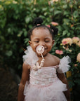 A young child in a pink dress smiles outdoors among greenery, holding a pacifier attached to the mushie Silicone Pacifier Clip | Luna.