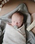 A baby sleeps peacefully in a crib, wrapped in a mushie Organic Cotton Muslin Swaddle Blanket with a pacifier. Nearby are a gray towel, a beige cream tube, and a soft pink cloth.