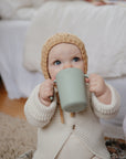 A baby in a knitted hat and cream sweater drinks from the Mushie Trainer Sippy Cup with easy-grip handles while sitting on a soft rug in a cozy room.
