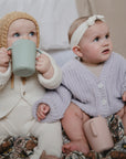 Two babies sit on a patterned blanket. One, in a knit hat and cream outfit, drinks from a green Mushie Trainer Sippy Cup. The other, wearing a headband and lavender sweater, holds a pink cup and gazes to the side.