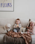Two babies sit on a white couch with a small white dog. One holds a mushie Lion Teether, while the other in a bunny-eared hat smiles with a drumstick. A brown blanket is on the couch, and a FLEURS poster hangs above.