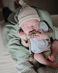 A newborn sleeps peacefully with a pacifier, wrapped in a green blanket on a striped cushion, wearing mushie’s Muslin Bib and a beige hat with a light onesie.