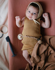 A baby in a mustard outfit and bonnet lies on the mushie Extra Soft Muslin Changing Pad Cover, surrounded by toys and a brown cloth, gazing up with wide eyes from the mauve pad.