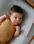 A baby in a mustard yellow sleeveless outfit lies on a mushie Extra Soft Muslin Changing Pad Cover next to a pink silicone spatula.