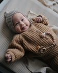 A baby in a cozy brown knit outfit and gray hat lies on the mushie Extra Soft Muslin Changing Pad Cover, smiling up with a pacifier on a beaded clip fastened to the sweater.