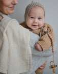 A smiling baby in a brown knit sweater and bonnet is held by an adult in a light gray top and cream shawl, wearing the mushie Silicone Pacifier Clip | Hera on their sweater and looking happily at the camera.