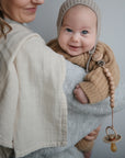 A smiling baby in a tan knit outfit holds a Mushie FRIGG Rope Natural Rubber Pacifier, clipped to their sweater. The baby's looking at the camera while being gently held, with the woman’s partial smile visible.