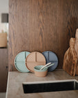 Three round silicone divided plates in pastel green, beige, and blue are propped against a wooden wall next to a beige bowl with two mushie Silicone Feeding Spoons. Wooden cutting boards stand nearby on the kitchen countertop.