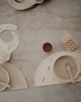 Round Dinnerware Plates by mushie on marble, with a dishwasher safe floral bib, silicone fork, placemat, cup, and phone-shaped toy. All crafted from non-toxic plastic in neutral pastel tones.