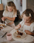Two young girls enjoy cake and cookies on mushie Round Dinnerware Plates. One girl is focused on her plate while the other stands nearby with dessert. A pink cup adds to the cozy, homey atmosphere.