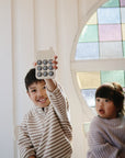 Two young children in striped sweaters sit indoors by a round stained glass window. The smiling boy holds up a mushie Phone Press Toy, designed to encourage fine motor skills, while the curious girl watches beside him.
