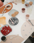 A white table set for a meal features mushie Square Dinnerware Plates, BPA-free and colorful, with plates holding raspberries, blueberries, crackers, and snack bowls. Cups and cutlery are included. A glass water bottle is also on the table.