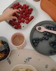A child’s hand reaches for raspberries on mushie Square Dinnerware Plates, with baby plates, blueberries, a pink cup, and utensils arranged on a white table.