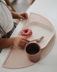 A young child sits at a table, reaching for a pink frosted donut with sprinkles on a Square Dinnerware Plate by mushie. A fork and cup are also present, all resting on a light pink placemat.