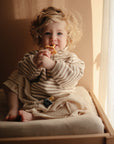 A young child with curly blonde hair sits on a mushie Extra Soft Muslin Changing Pad Cover by a sunlit window, wearing a striped beige outfit and holding a yellow teething toy, with a pacifier resting nearby.