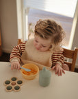 A young child practices fine motor skills while eating fruit, with a mushie Flower Press Toy and a drink bottle on the table. Light streams in from a window behind them.
