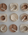 A grid of nine mushie Square Dinnerware Plates, bowls, and utensils in beige and brown, neatly arranged on a light surface. Each BPA-free set features minimalist square plates with matching forks or spoons.