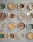 A flat lay of fifteen mushie Square Dinnerware Plates, BPA-free and pastel-colored in beige, green, brown, and yellow, each set with matching spoons or forks displayed on a light background.