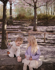 Two young girls sit under blossoming trees, happily using mushie Snack Cups. Pink petals surround them, creating a peaceful spring setting perfect for toddlers enjoying their spill-proof silicone cups.
