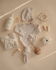 A flat lay of neutral baby items on a beige background, featuring the mushie Ribbed Baby Bonnet, a knotted onesie, swaddle, bottle, lotion, rattle, pacifier, teether, and cup—all in soft muted tones.
