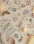 A flat lay of mushie baby accessories, including the Flower Training Toothbrush and pastel-colored teethers, rattles, stacking toys, pacifiers, wooden beads, rings, and a rainbow, arranged on a cream surface with small daisy decorations.