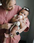 A woman in a rose-colored robe holds a baby wearing a bow headband and patterned outfit. The baby chews on an egg-shaped teether attached to the mushie Silicone Pacifier Clip | Hera, featuring a minimalist design, while the woman supports her and smiles.